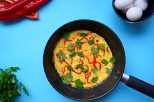 Kuvassa on paistinpannulla paprikafrittata. Kuvan tausta on sininen.