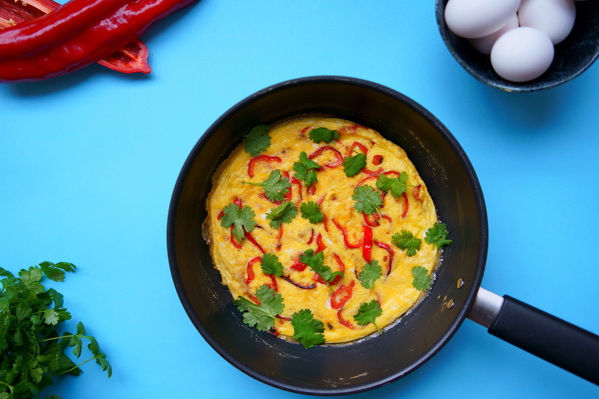 Kuvassa on paistinpannulla paprikafrittata. Kuvan tausta on sininen.